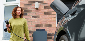 A woman at a residential location approaching an electric vehicle with a charging cable in hand, ready to plug it in.