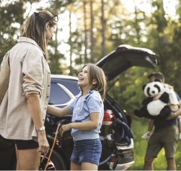 Mother and daughter standing beside an electric vehicle being charged whilst father unloads second child and large panda soft toy from vehicle